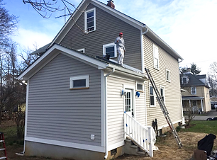 A person on a ladder repairs the roof of a two-story house with gray siding.